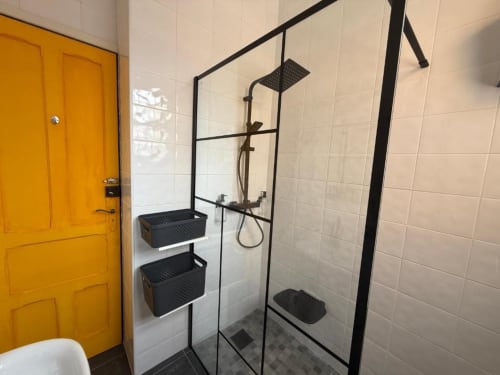 Banheiro moderno com box de vidro de estrutura preta, azulejos brancos, cestos de armazenamento pretos e porta amarela