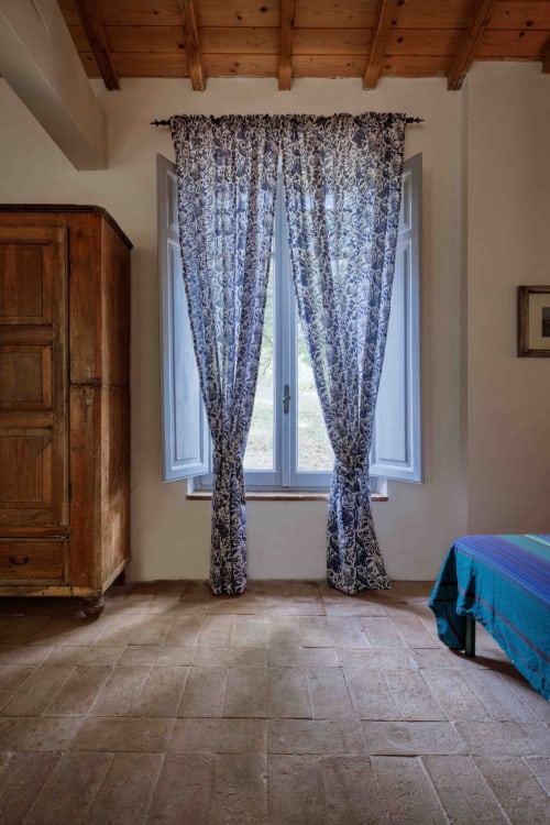 Ampia camera con tende floreali blu alle finestre alte, soffitto con travi in legno, pavimenti in cotto e mobili in legno