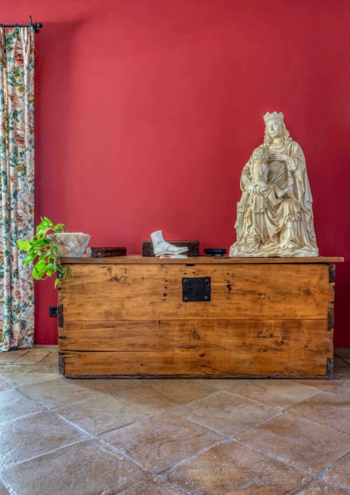 Baule in legno rustico con statua classica e pianta in vaso davanti a un muro rosso vivace nella fattoria Donkeys & Roses, Viterbo