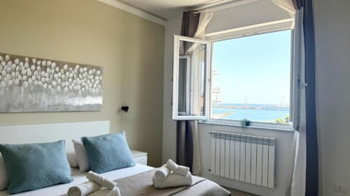 Camera da letto luminosa a Palermo con vista mare, cuscini blu, testata bianca e porto visibile dalla finestra aperta