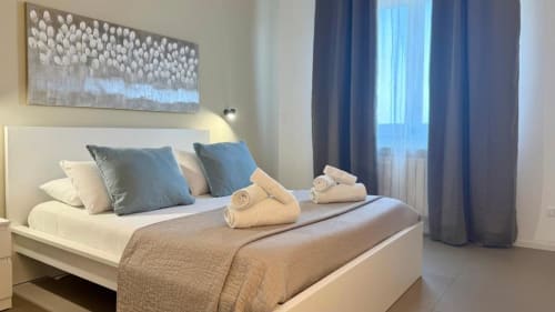 Camera da letto moderna a Palermo con vista sul mare, letto bianco, cuscini blu, asciugamani arrotolati e finestre con affaccio sull'oceano