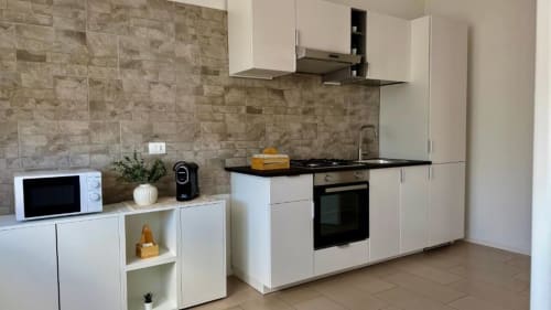 Cucina moderna bianca nell'appartamento di Palermo con piano cottura nero, forno incassato, rivestimento in pietra e microonde.
