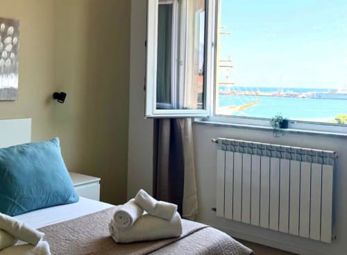 Luminosa camera da letto a Palermo con vista mare, cuscino blu, asciugamani bianchi, termosifone e porto turchese visibile
