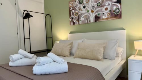 Camera da letto moderna nell'appartamento palermitano con letto bianco, asciugamani arrotolati, cuscini a righe, pareti verdi, lampada nera e stampa astratta