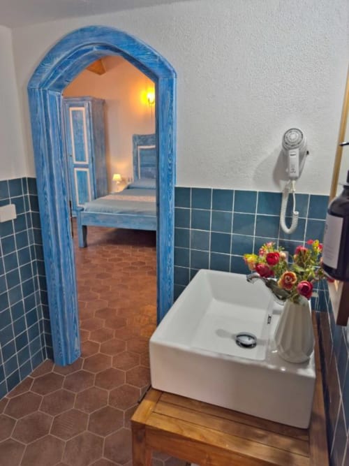 Bagno con lavabo bianco, fiori freschi e arcata azzurra verso la camera nella suite boutique di Bosa