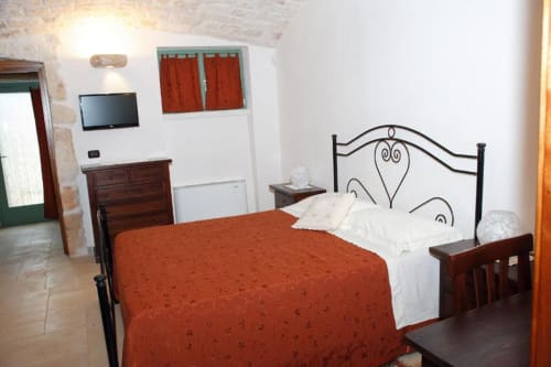 Spaziosa camera da letto in Trulli Boccadoro con letto in ferro battuto, coperta rossa, cuscini bianchi, cassettone in legno, TV a schermo piatto e