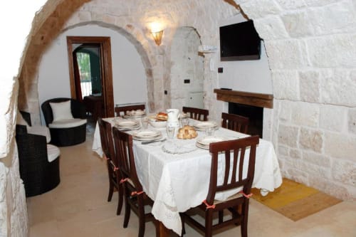 Sala da pranzo in pietra nel Trulli Boccadoro con tavolo con tovaglia bianca, sedie in legno e soffitto ad arco