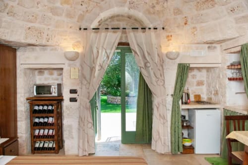 Interno accogliente di trullo con volta in pietra, porta verde verso il giardino, tende beige, cantina vinicola, cucina e