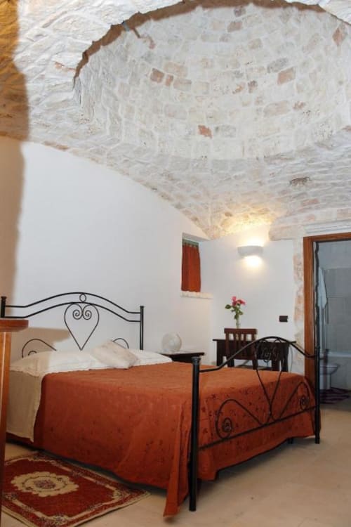 Camera da letto rustica con soffitto in pietra a vista, letto in ferro battuto con coperta rossa, muri bianchi e tradizionali Trulli