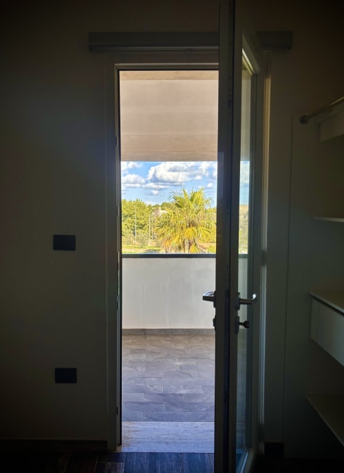 Porta in vetro aperta della B&B Hélios a Nardò che rivela un balcone con palma e vista sulla campagna
