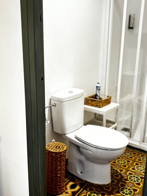 Compact bathroom at Mas Ribas Hotel in Palamos with white toilet, decorative yellow patterned floor, and woven storage basket