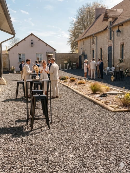 Réception de mariage dans la cour du Domaine du Grand Fort avec des invités vêtus de crème près des tables et des bâtiments en pierre en Normandie