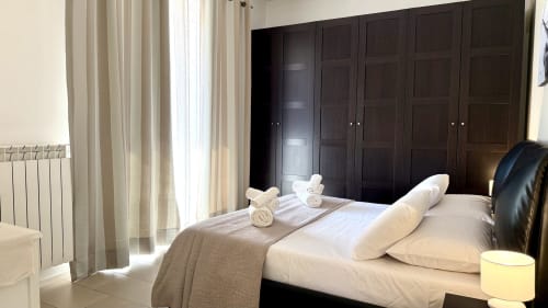 Camera da letto moderna nell'appartamento di Palermo con biancheria bianca, armadio in legno scuro e vista sulla città dalle porte della terrazza