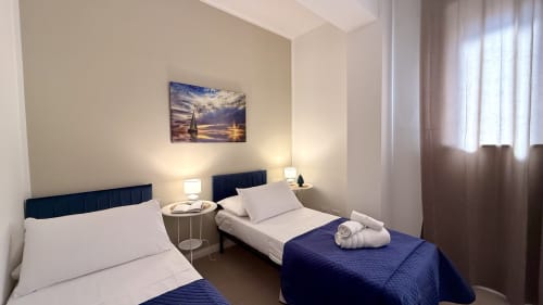 Camera da letto doppia a Palermo con testiere blu scuro, biancheria bianca, asciugamani arrotolati, quadro a vela e luce naturale