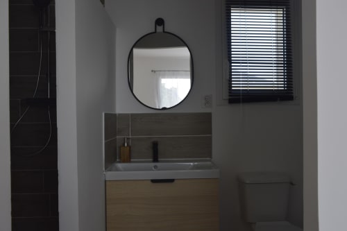 Salle de bains moderne avec miroir ovale, vasque en bois et douche aux carrelages foncés du gîte Résidence des dunes à Landéda