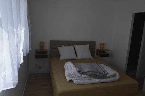 Chambre à la Résidence des dunes à Landéda avec lit, oreillers, tables de nuit et fenêtre aux voilages délicats