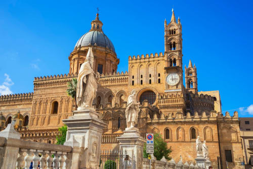 Cattedrale di Palermo con cupola dorata e campanile sotto cielo azzurro, con statue in marmo in primo piano