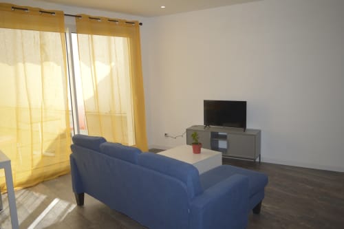 Salon moderne du gîte de Landéda avec canapé bleu, téléviseur, table basse blanche et rideaux jaunes donnant sur la piscine