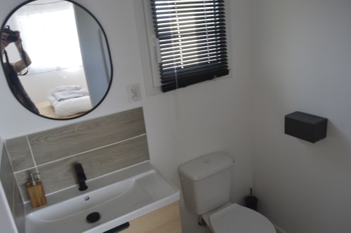 Salle de bain moderne avec miroir rond, vasque en bois, toilettes et fenêtre au gîte Résidence des dunes à Landéda