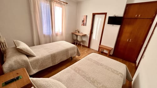 Two twin beds in a bright hostel room at Hostal Estació in Puigcerdà with wooden wardrobe, desk, TV and mountain views