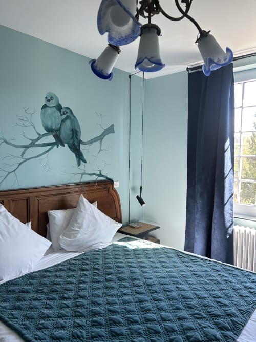 Charming guest room at Clos d'Azel with blue glass paste pendant lights, hand-painted bird mural, hotel-quality bed and garden view in Alsace