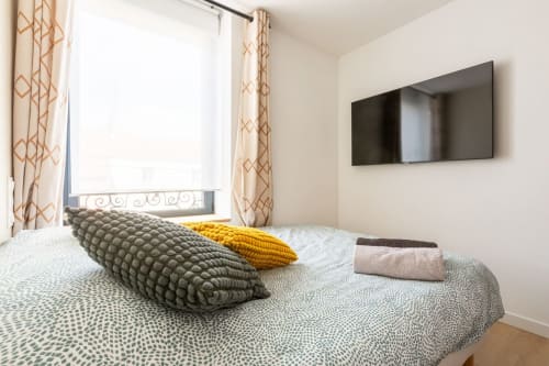Chambre moderne de l'appartement Le Cocon à Angres avec lit, coussins décorés, télévision murale et rideaux à motifs