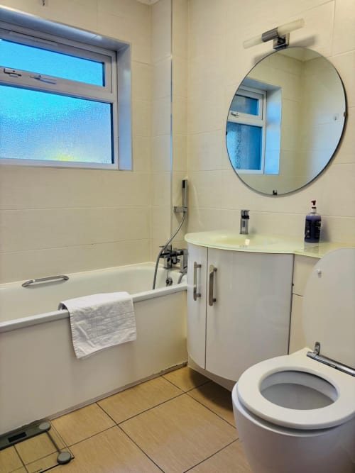 Modern bathroom in England Furnished Homes, High Wycombe with bathtub, white vanity, circular mirror, toilet, and window.