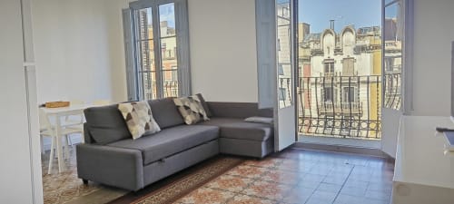 Sala de estar de apartamento moderno en Barcelona con sofá seccional gris, balcón con vistas al paisaje urbano del distrito de Eixample, y