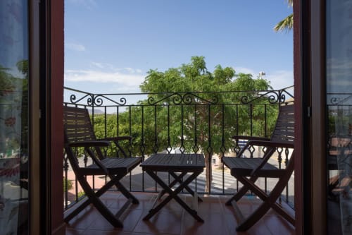 Sunny balcony with two black chairs and small tables overlooking green trees and city views at Alojamientos Carolina y Vanesa