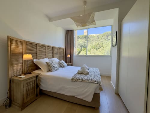 Chambre lumineuse et moderne dans l'appartement en location Eugénie à Ajaccio en Corse avec tête de lit en bois, literie blanche et baies vitrées offrant une vue panoramique sur la mer