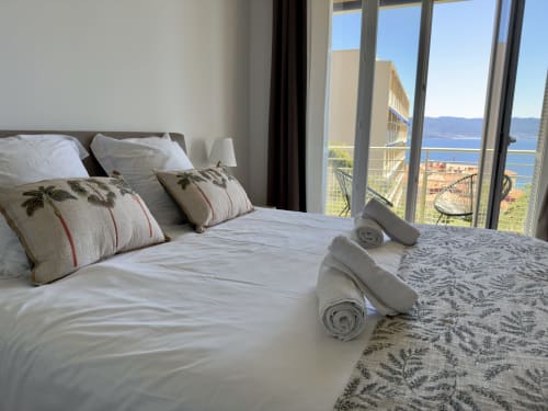 Chambre lumineuse dans l'appartement en location Eugénie à Ajaccio en Corse avec balcon baies vitrées offrant une vue panoramique sur la mer, lit aux coussins décorés et serviettes roulées