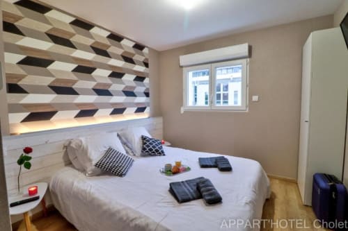 Chambre moderne de l'Appart'Hôtel Cholet avec tête de lit géométrique, literie blanche, coussins à motifs et vue sur le jardin.