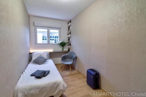 Chambre moderne à l'Appart'Hôtel Cholet avec lit simple, fauteuil, bureau, fenêtre sur jardin et parquet
