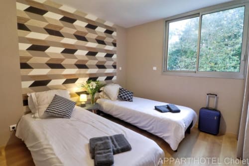Chambre twin à l'Appart'Hôtel Cholet avec tête de lit géométrique en bois, literie blanche, coussins à carreaux noirs et vue sur le jardin