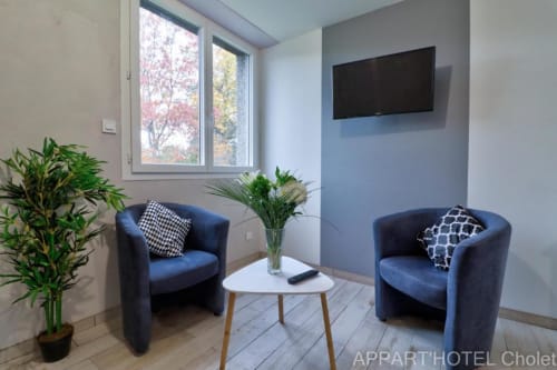 Coin lounge moderne à l'Appart'Hôtel Cholet avec deux fauteuils bleus, table blanche, TV écran plat, plantes en pot et jardin