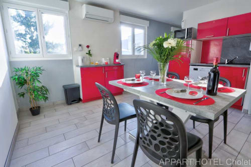 Espace cuisine-séjour moderne de l'Appart'Hôtel Cholet avec armoires rouges, comptoirs blancs et mobilier contemporain.