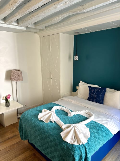 Acogedora habitación de apartamento parisino con pared de acento verde azulado, vigas de madera, armario blanco y ropa de cama color turquesa cerca del Louvre