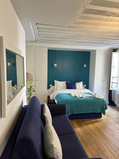 Recámara de apartamento parisino moderno con pared de acento verde azulado, muebles crema, vigas de madera y sofá azul marino cerca del Louvre