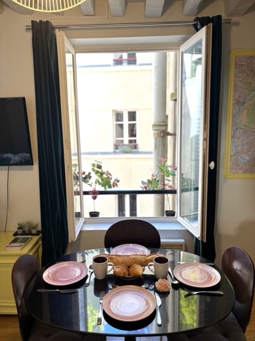 Elegante comedor en apartamento parisino con mesa redonda negra, platos rosas, cruasanes y puertas francesas al jardín