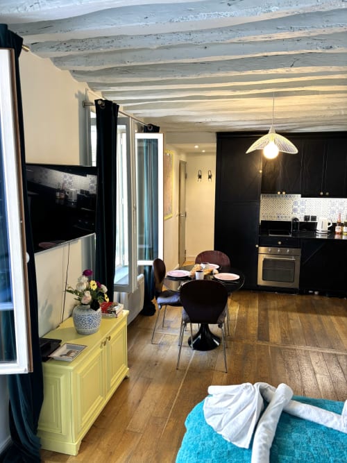 Acogedora cocina-comedor de apartamento parisino con gabinetes negros, vigas de madera, cómoda amarilla y vistas al jardín