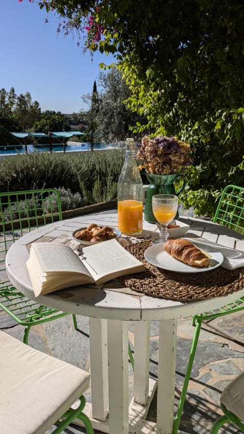 Breakfast setting at Casas de Juromenha with book, croissant, juice overlooking pool and gardens