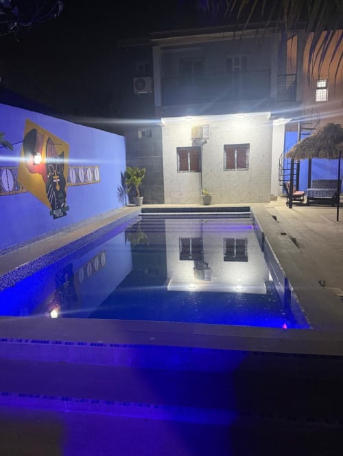 Cour de l'hôtel Keur Medzo la nuit avec piscine illuminée en bleu, bâtiment blanc et murs décorés à Lac Rose, Niaga