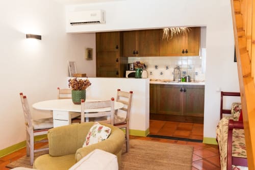 Aconchegante área de cozinha-sala na villa Casas de Juromenha com mesa de jantar, poltrona amarela e móveis de madeira