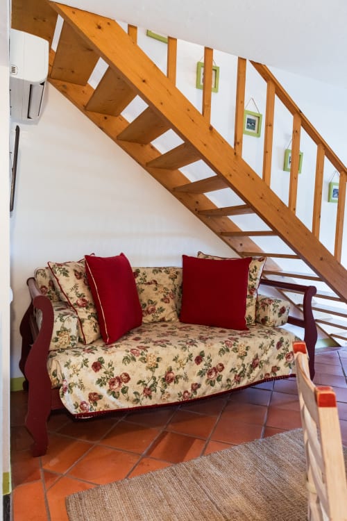 Aconchegante área de estar sob a escada de madeira na villa Casas de Juromenha com sofá floral e almofadas vermelhas