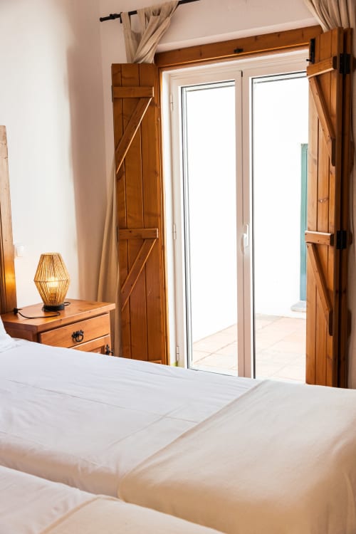 Quarto brilhante com roupa de cama branca, portas de celeiro em madeira, luminária de vime e portas de vidro que abrem para o terraço em Casas de Juromenha