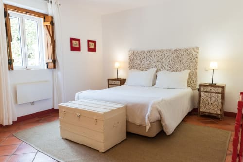 Quarto ensolarado na villa Casas de Juromenha com cama branca, cabeceira estampada, mesinhas de madeira e cômoda creme