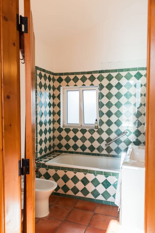 Banheiro com padrão de azulejos em diamante verde e branco, banheira, sanitário e janela na villa Casas de Juromenha em Portugal