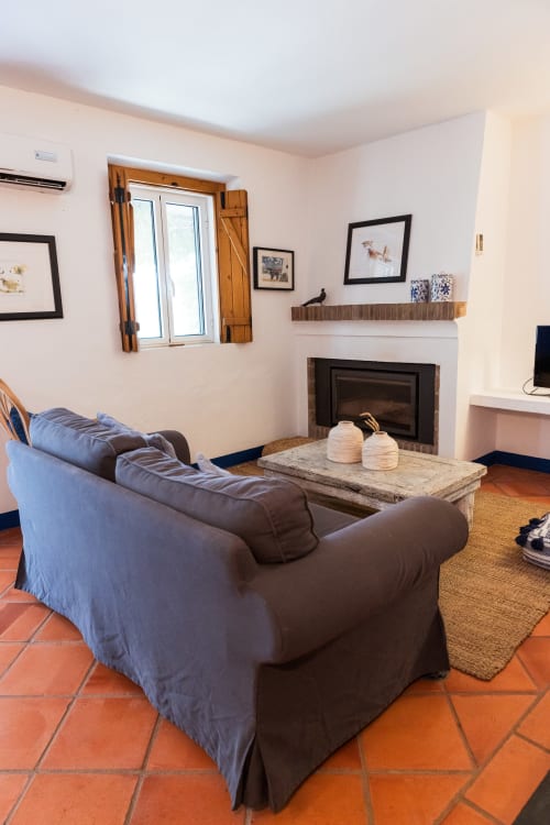 Sala de estar aconchegante na villa Casas de Juromenha com lareira, sofá azul, azulejos de terracota e janela com vista para o jardim