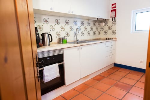 Cozinha branca moderna com azulejos florais no respaldo, fogão preto, pia e piso de ladrilho terracota na villa Casas de Juromenha