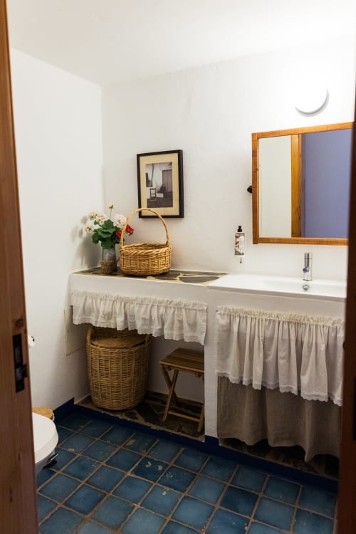 Banheiro rústico na villa Casas de Juromenha com espelho de moldura de madeira, pia branca, cestos de vime e piso de azulejos azuis.